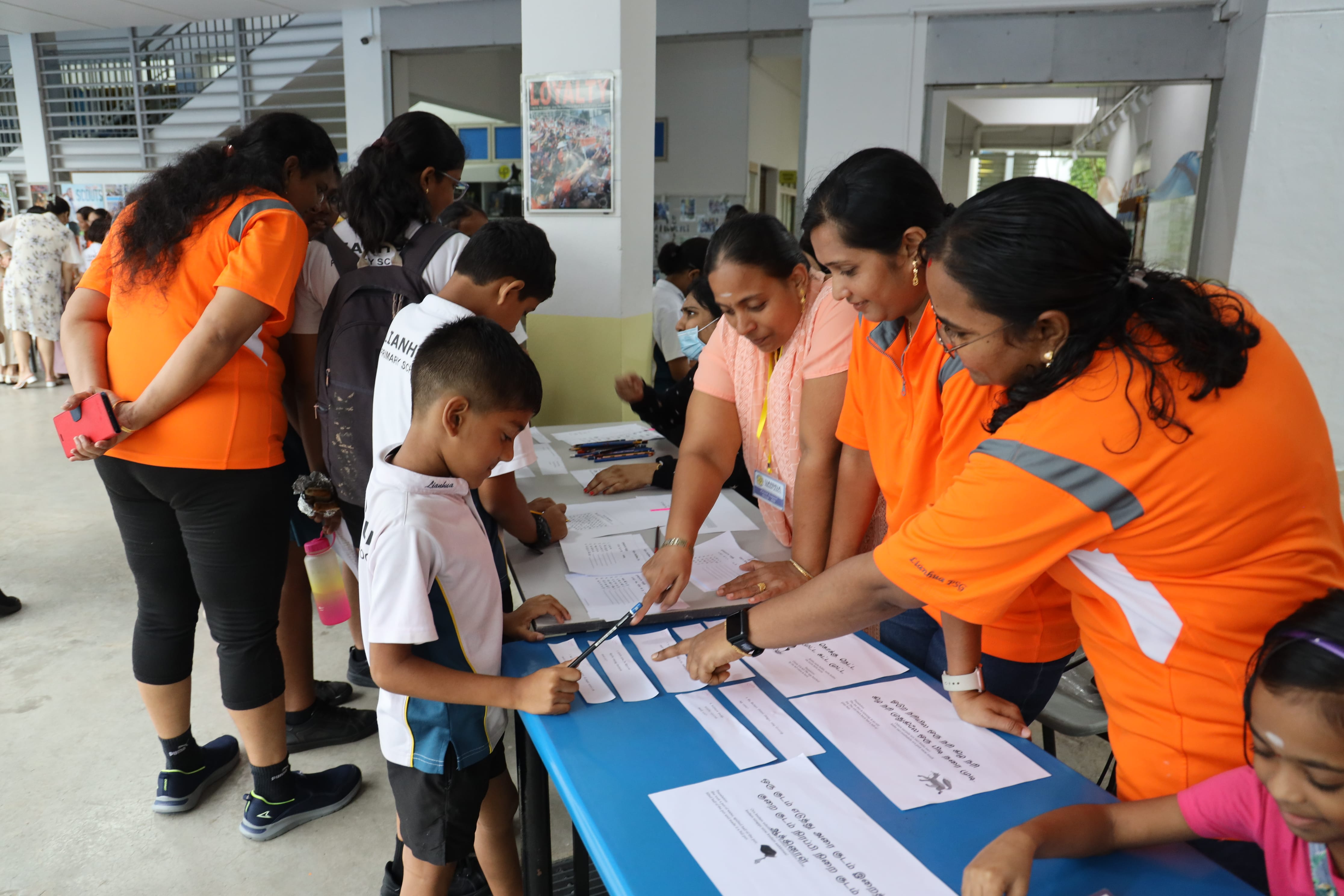 Women in orange shirts guide students signing papers at a table in a school corridor.